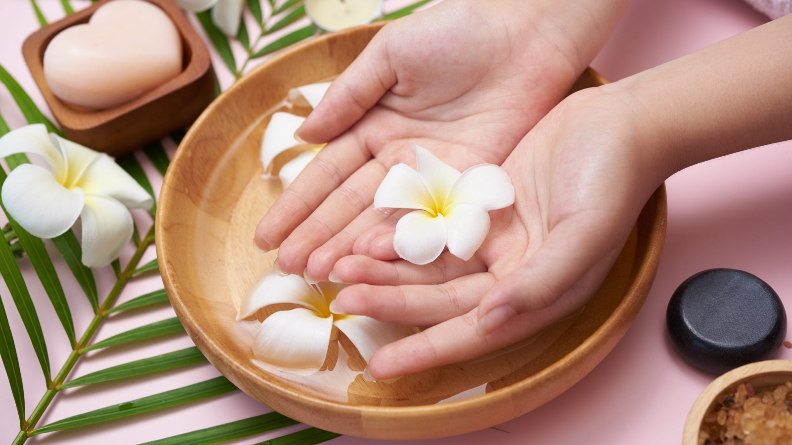 Woman soaking her hands in bowl of water and flowers, Spa treatment and product for female feet and hand spa, massage pebble, perfumed flowers water and candles, Relaxation. Flat lay. top view.